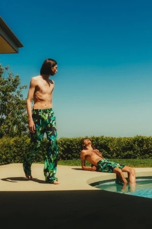 Two shirtless men in tropical-print bottoms relax poolside in bright daylight under a clear blue sky surrounded by green foliage.