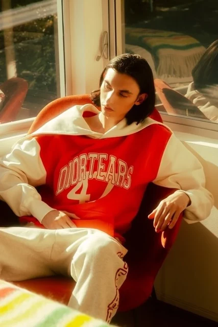 Young adult man sits in a red chair by a window, wearing a white and red Dior Tears hoodie and sweatpants in soft, diffused sunlight.