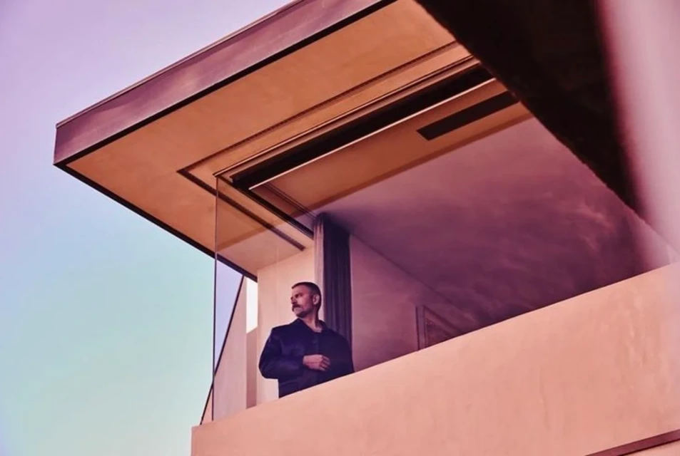 A man stands on a modern building's balcony with a glass railing, wearing a jacket, in front of a pink and blue sky.
