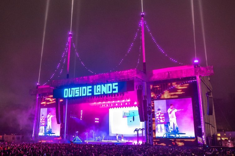 The music festival 'Outside Lands' is captured at night, showcasing a large crowd in front of a stage with large video screens and light beams.