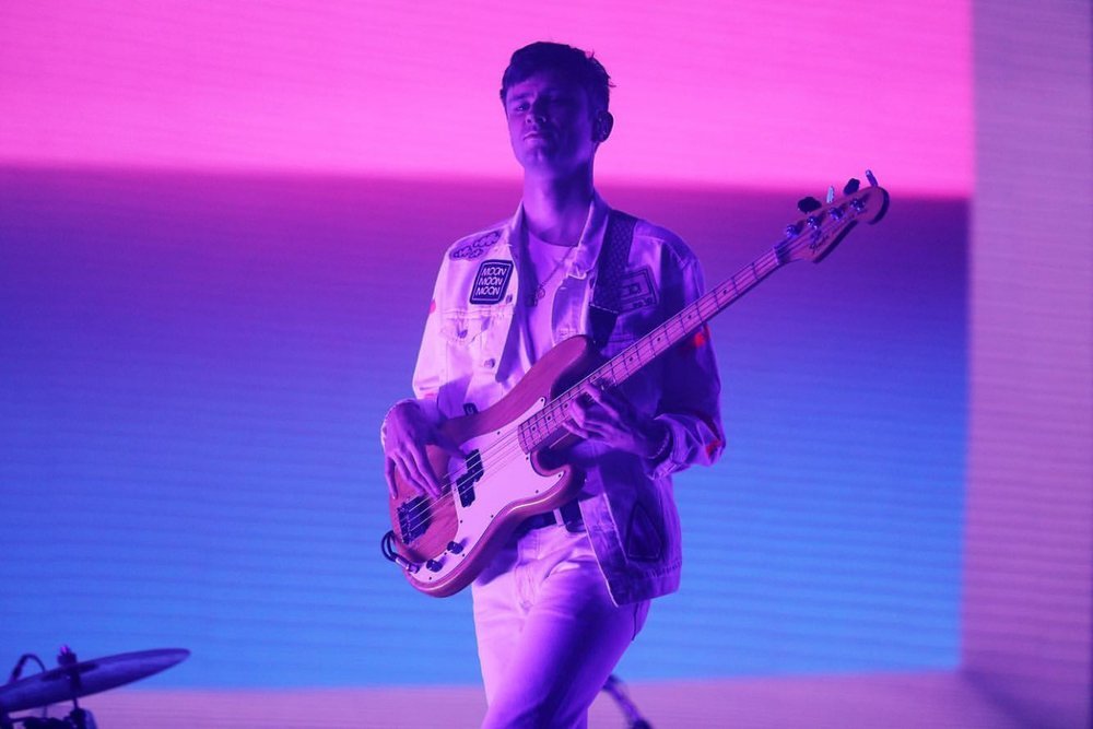 Male musician playing bass guitar on stage, bathed in blue and pink light, wearing white outfit with patches on jacket.