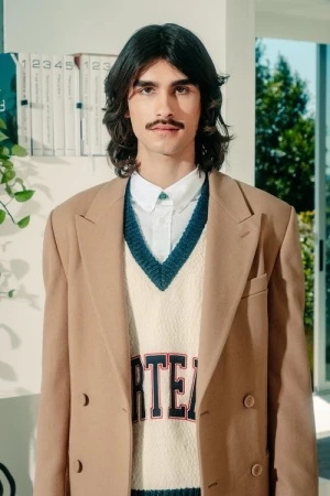 A man with long dark hair and a mustache wearing a beige blazer over a white shirt and sweater vest, styled by Kenn Law.