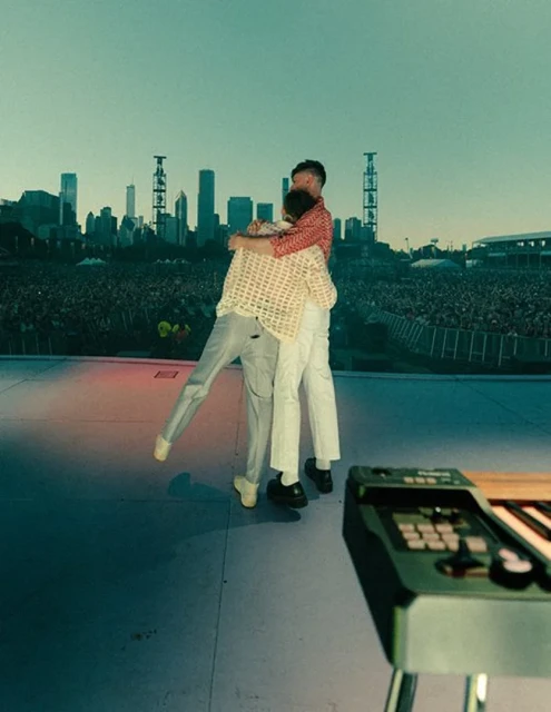 Two adult men embracing on a stage in front of a large crowd and city skyline backdrop, with a keyboard visible in the foreground.