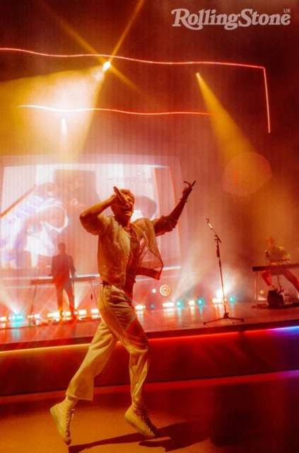 Male musician performs on stage in white outfit with keyboardist and backing musician at concert with orange stage lighting.