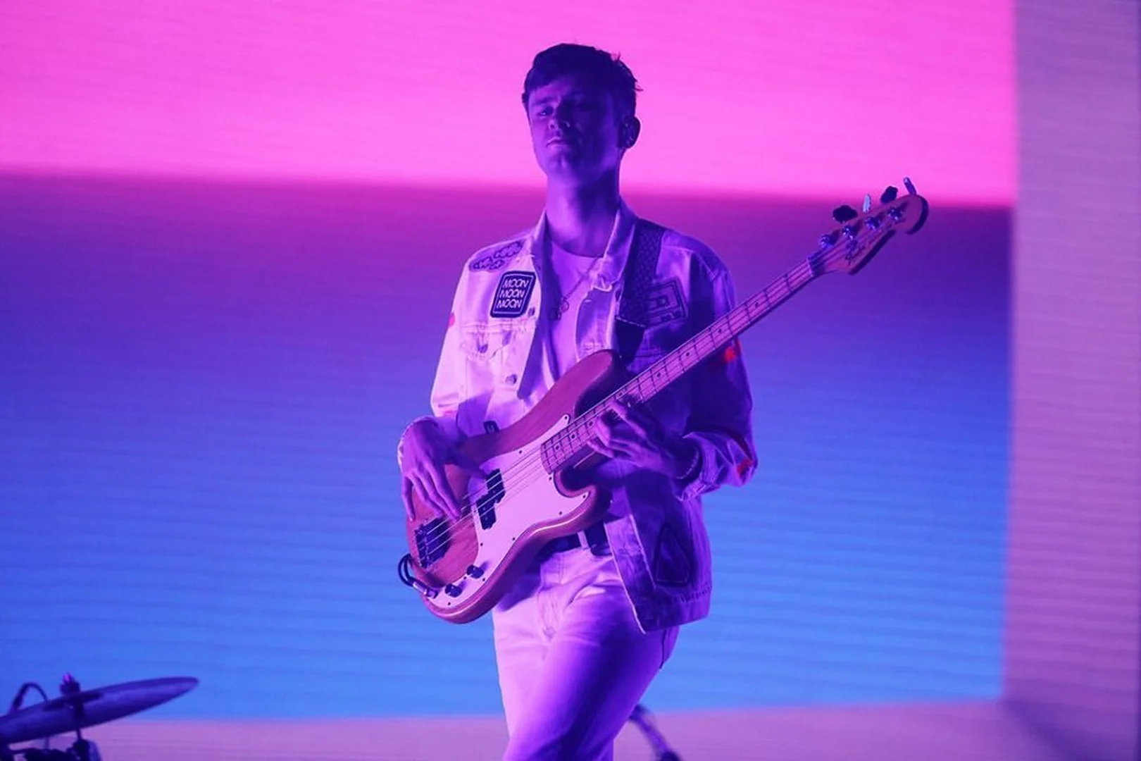 A person plays an electric bass guitar illuminated by blue and pink stage lighting, wearing a white denim jacket.
