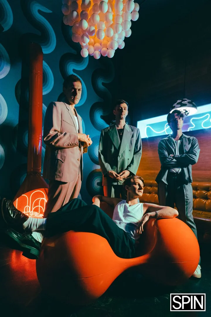 Stylish group portrait of four men in suits in a modern, retro-styled indoor setting with unique lighting and bold color palette.