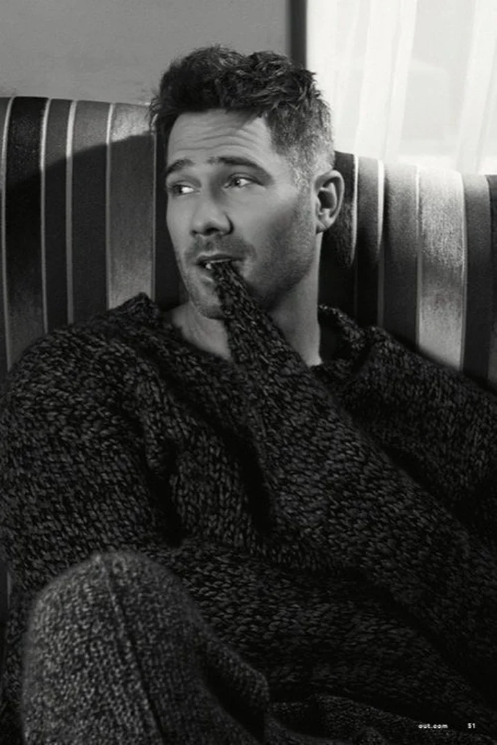 Portrait of an adult male with dark hair wearing a sweater, sitting on a striped couch indoors, looking left with a finger in his mouth.