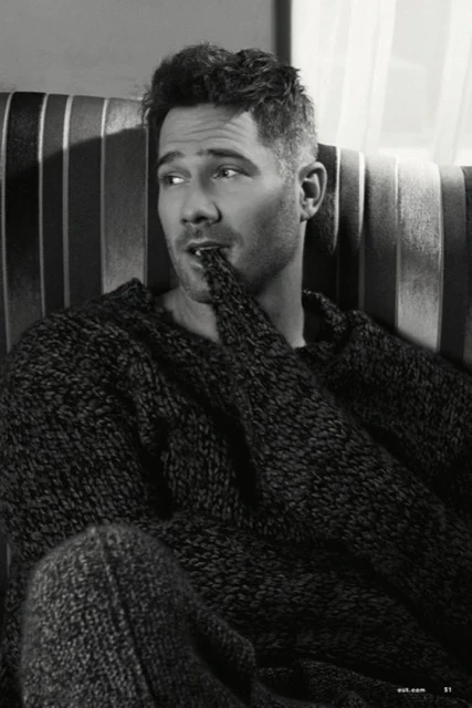 Portrait of an adult male with dark hair wearing a sweater, sitting on a striped couch indoors, looking left with a finger in his mouth.
