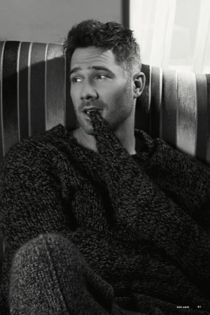 Portrait of an adult male with dark hair wearing a sweater, sitting on a striped couch indoors, looking left with a finger in his mouth.