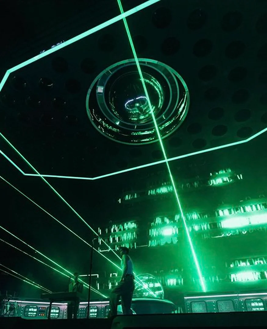 A futuristic stage bathed in green light features two figures standing on a platform, with geometric patterns and shapes in the background.
