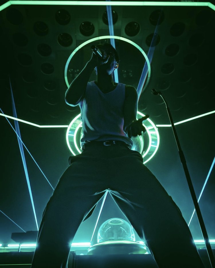 Low angle view of a man singing into a microphone on stage with neon lights. He is wearing a tank top and trousers.