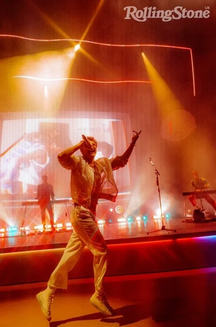 Singer performs on stage in bright orange lighting during live concert with band, wearing sheer sparkling top and white pants.