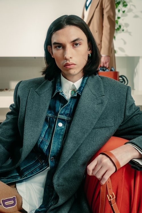 Stylish man in gray blazer and denim jacket poses indoors, holding brown leather bag. Another man and potted plant visible in background.