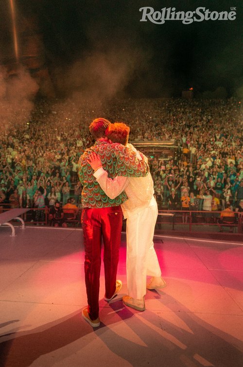 Two men embracing on stage in front of a crowd, warmly lit by stage lights; crowd in the background under dark sky.