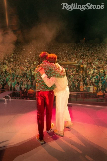 Two men embracing on stage in front of a crowd, warmly lit by stage lights; crowd in the background under dark sky.