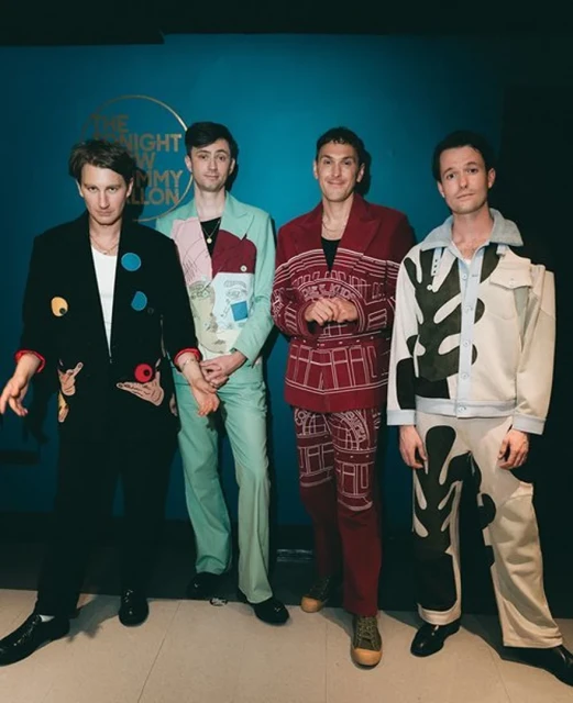 Four people stand in a row against a blue wall with a logo, wearing patterned jackets and pants with black shoes and smiling subtly.