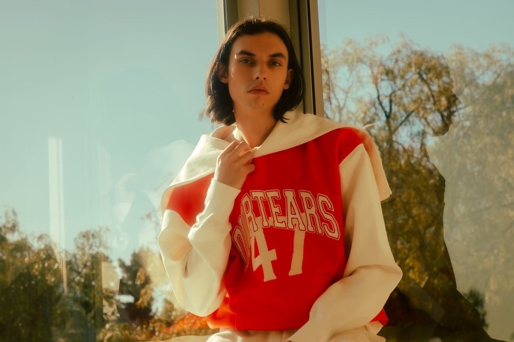 Young man with dark hair wearing a red and white varsity sweatshirt with lettering against a large glass window.