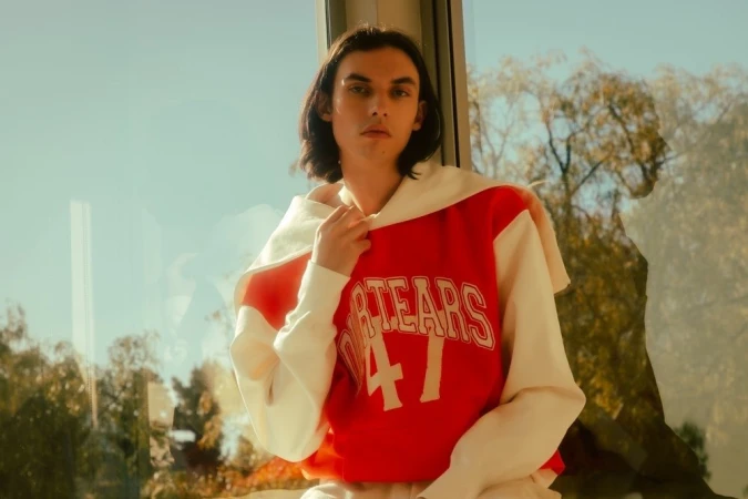 Young man with dark hair wearing a red and white varsity sweatshirt with lettering against a large glass window.