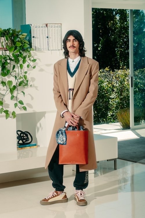 A man in a tan coat and orange tote bag stands in a modern interior with plants and bright lighting, capturing a contemporary fashion look.