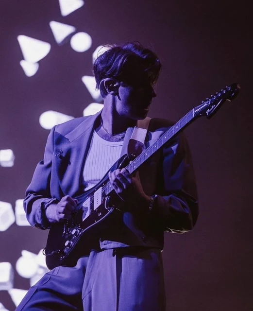 A person playing an electric guitar on stage, wearing a suit under dark lighting with geometric light patterns in the background.