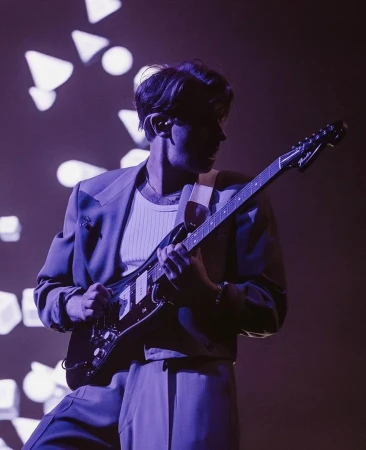 A person playing an electric guitar on stage, wearing a suit under dark lighting with geometric light patterns in the background.