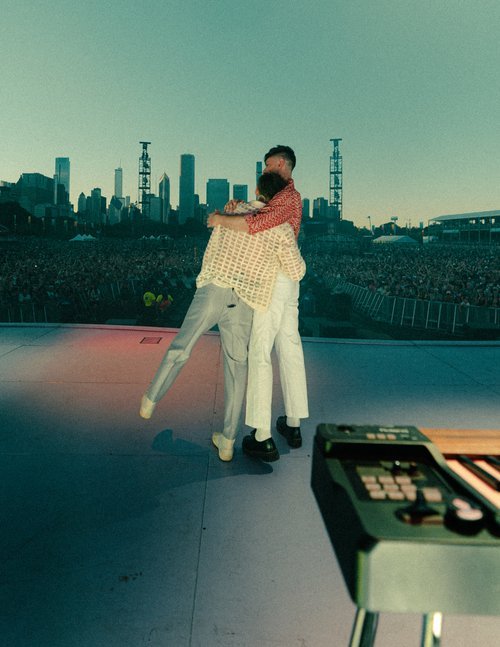 Two people embrace on a stage in front of a large crowd and city skyline, with a keyboard in the foreground, in muted colors.