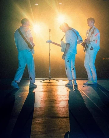 Three musicians on stage in matching outfits backlit by bright stage lights, playing guitars, captured in a medium shot.