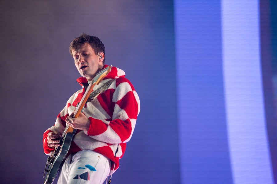 A musician sings and plays an electric guitar. The singer wears a red and white checkerboard fleece jacket and white pants on stage.