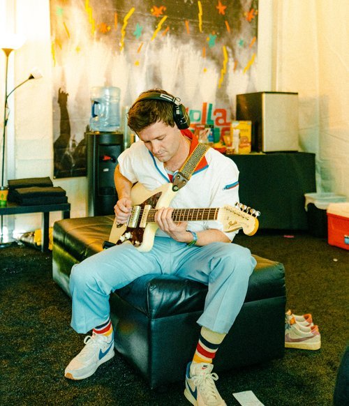 Young adult male musician with headphones plays electric guitar indoors sitting on a black ottoman in front of a colorful backdrop.