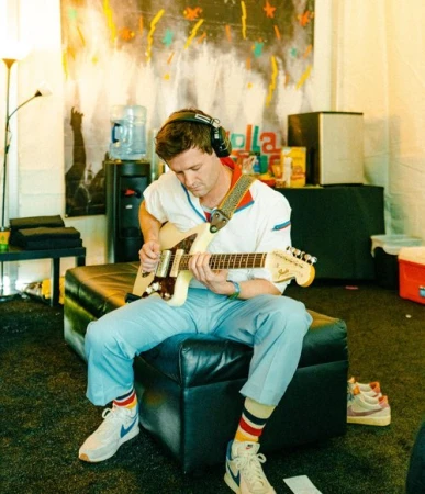 Young adult male musician with headphones plays electric guitar indoors sitting on a black ottoman in front of a colorful backdrop.