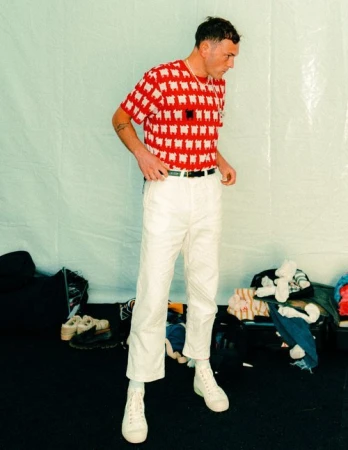 Man wearing a red and white patterned t-shirt and white trousers standing against a pale background