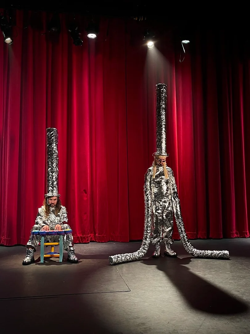 Two figures in reflective costumes on a stage with red curtains and spotlight, one seated at a xylophone, the other standing.