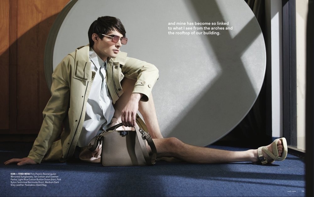 Man poses in fashion editorial wearing sunglasses, parka, button-down shirt, shorts, and sandals, seated next to a grey leather bag.