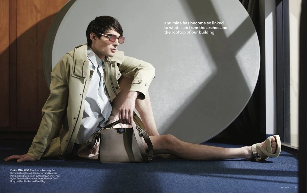 Man poses in fashion editorial wearing sunglasses, parka, button-down shirt, shorts, and sandals, seated next to a grey leather bag.