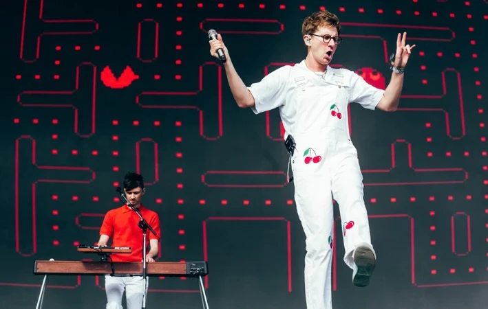 Singer wearing a white jumpsuit performing on stage with a bandmate playing keyboard against a Pac-Man themed background.