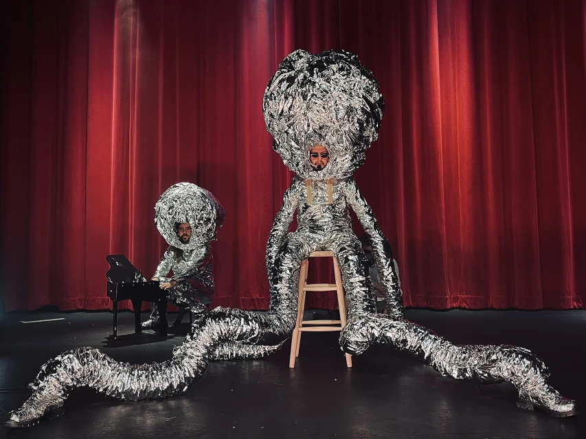 Two performers in shiny foil costumes on stage, one plays a black piano, the other sits on a stool, a red curtain in the background.