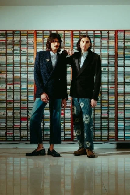 Two men pose in blazers and patterned pants against a backdrop of stacked audio cassettes, creating a vintage and stylish visual.