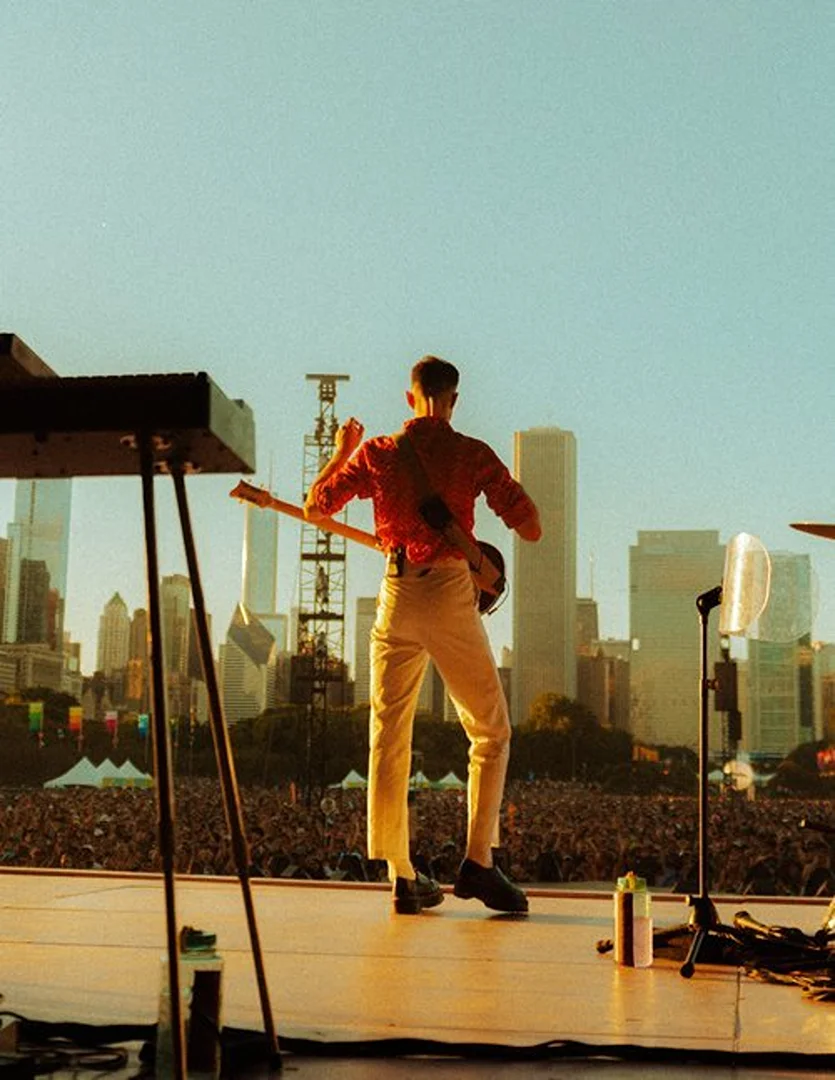 Back view of a musician performing on stage in front of a large crowd and city skyline, illuminated by warm stage lights.