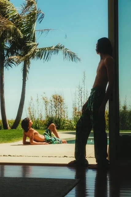 Two shirtless young men in patterned pants and shorts relax outdoors on a sunny day with palm trees and a swimming pool in the background.