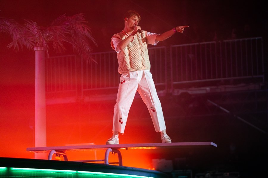 Male singer performs on a stage with a microphone, wearing a knit vest and white pants with cherry designs. Bright red stage lighting.