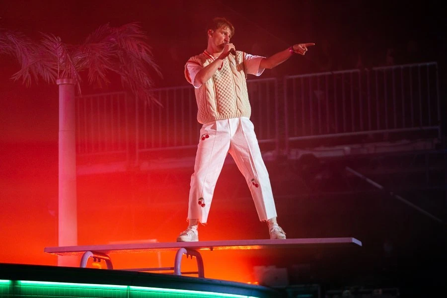 Male singer performs on a stage with a microphone, wearing a knit vest and white pants with cherry designs. Bright red stage lighting.
