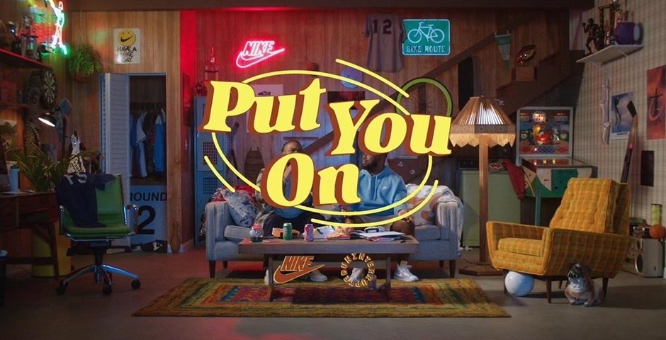 Two men relax on a couch in a retro-styled room with neon signs and vintage decor, promoting the slogan 'Put You On'.