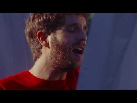 Man in a red shirt sings with mouth open and eyes closed in front of a blurry blue background, medium close-up shot.