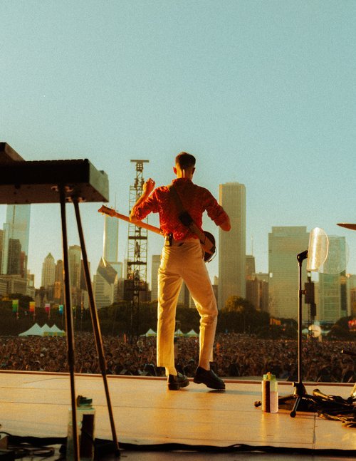 Performer on stage with guitar faces audience, city skyline in background at outdoor concert during golden hour lighting.