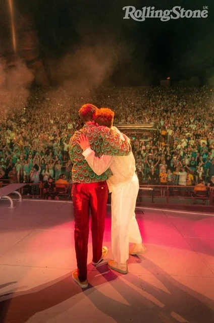 Two men embrace on stage in front of large crowd at a music concert, bathed in orange and red lighting with Rolling Stone logo visible.