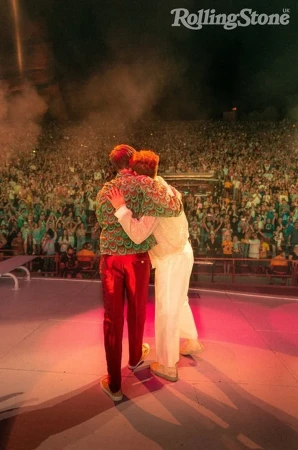 Two men embrace on stage in front of large crowd at a music concert, bathed in orange and red lighting with Rolling Stone logo visible.