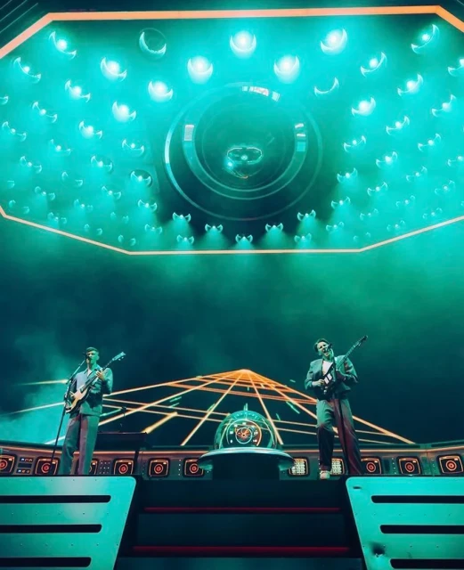 Two guitarists perform on a futuristic stage with teal lighting and geometric backdrop. The lighting emphasizes the setting and musicians.