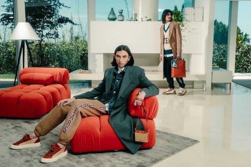 Two adult male fashion models pose indoors in stylish loungewear and outerwear with a modern interior and natural light from large windows.