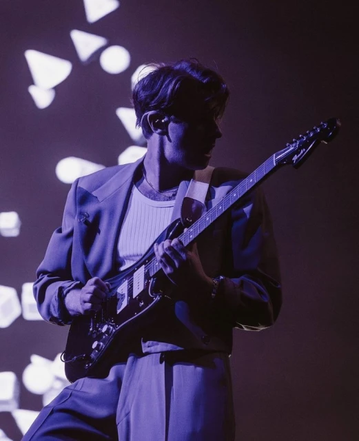 Musician playing electric guitar on stage with geometric light backdrop, wearing a suit in a dimly lit, music venue performance.