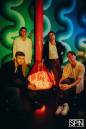 Four men posed around a red neon fireplace against a vibrant green and blue retro patterned wall.